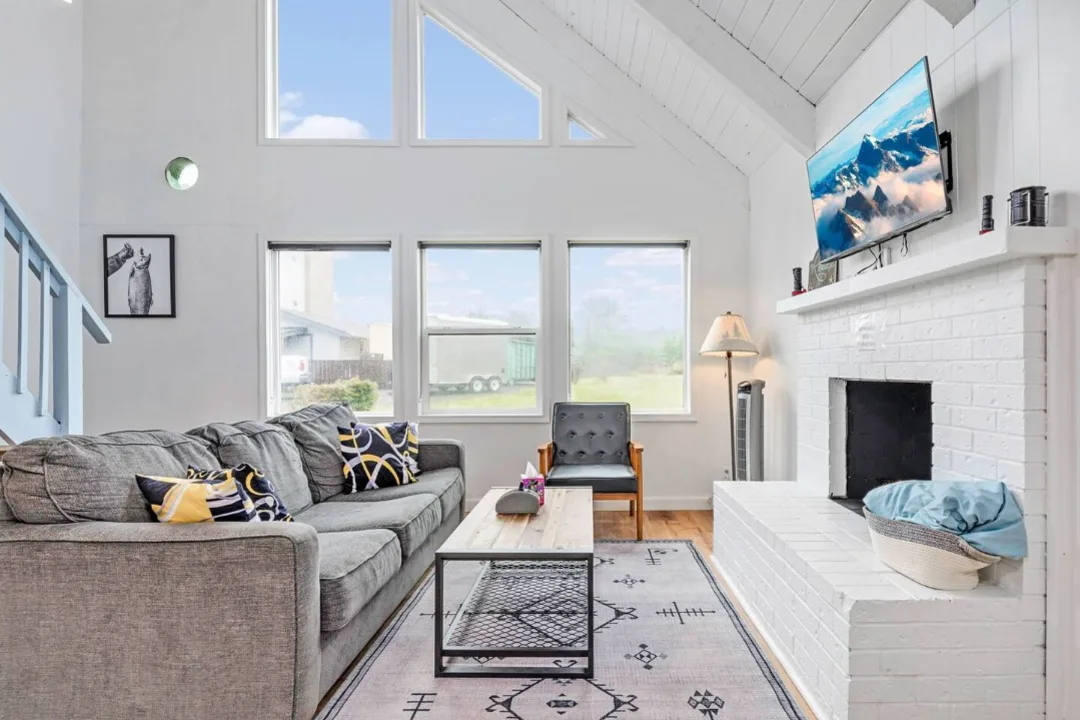 Living room of Tidal House Chalet 2 with a gray sofa beneath a vaulted white ceiling and beside a white fireplace.