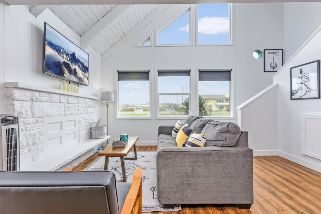 Vaulted living room of Tidal House Chalet 9 with a gray sofa, white fireplace, and coastal artwork.